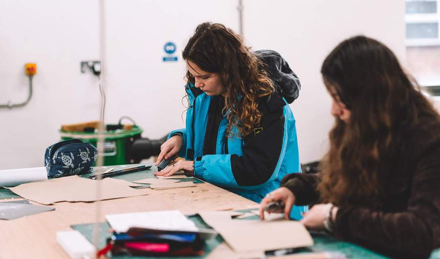 Students cutting cardboard pieces on a workshop table with craft tools and materials spread out.