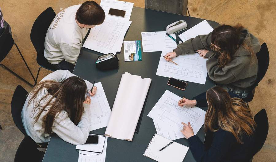 Group of students seated around a table studying architectural plans with notebooks and phones nearby.