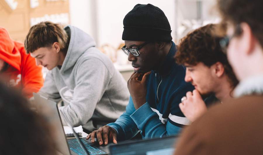 Students seated at a table working on open laptop computers in a studio environment.