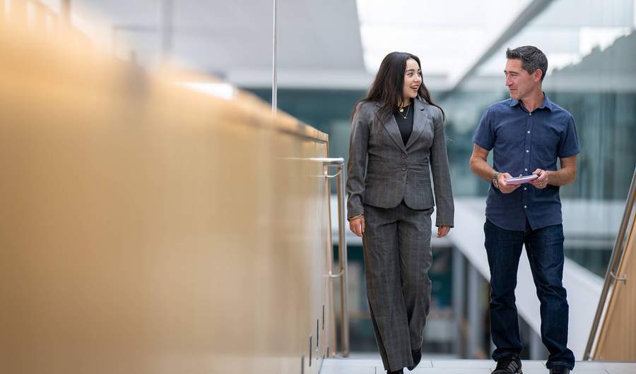 Two people walk alongside each other at the top of a staircase. In the background are glass walls overlooking an open atrium space.