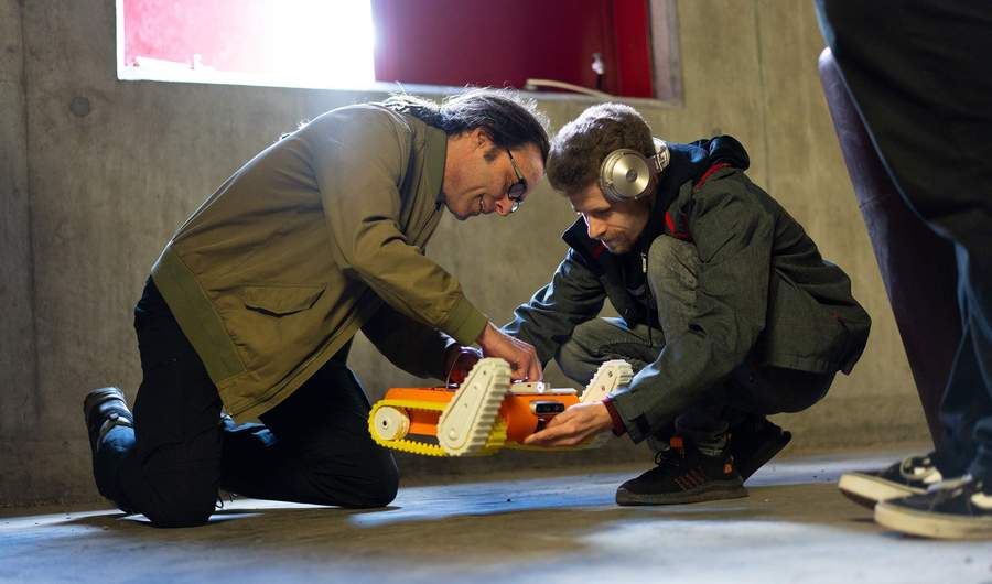 A person holds a small robot with caterpillar tracks while another person adjusts wires on its casing. They are in a room with a bare concrete floor and walls.