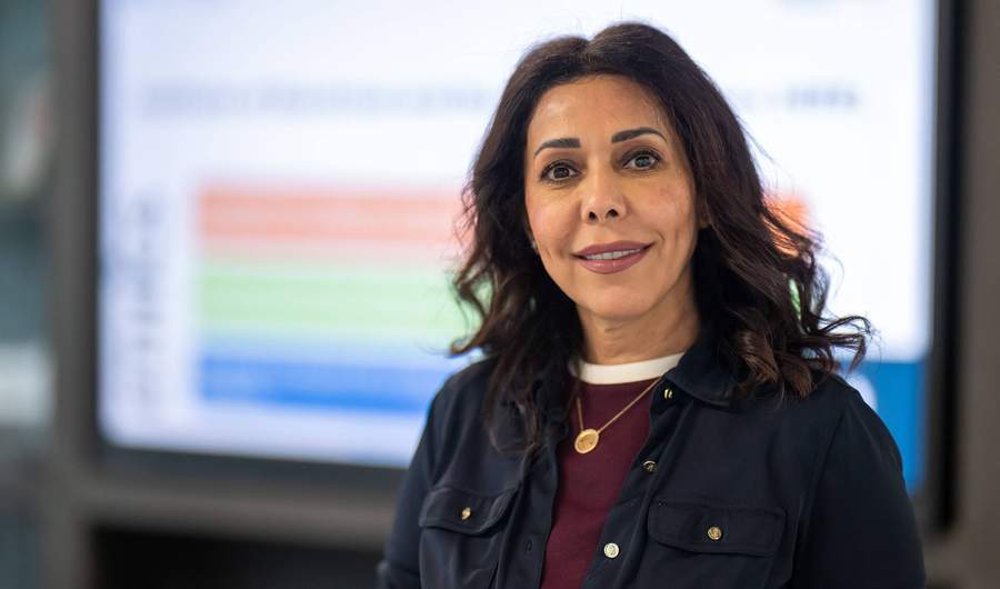 A woman wearing a dark blue jacket and maroon sweater. In the background is a large display screen.