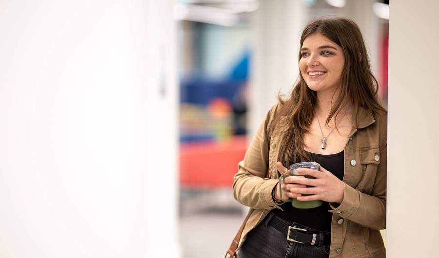 A young woman leaning against a wall, holding a reusable coffee mug.