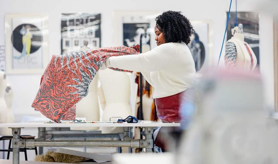 A person with curly hair holds up a colourful fabric in a design studio. In the background, a mannequin and framed art are visible. The room is well-lit, creating an inspiring workspace for creativity.