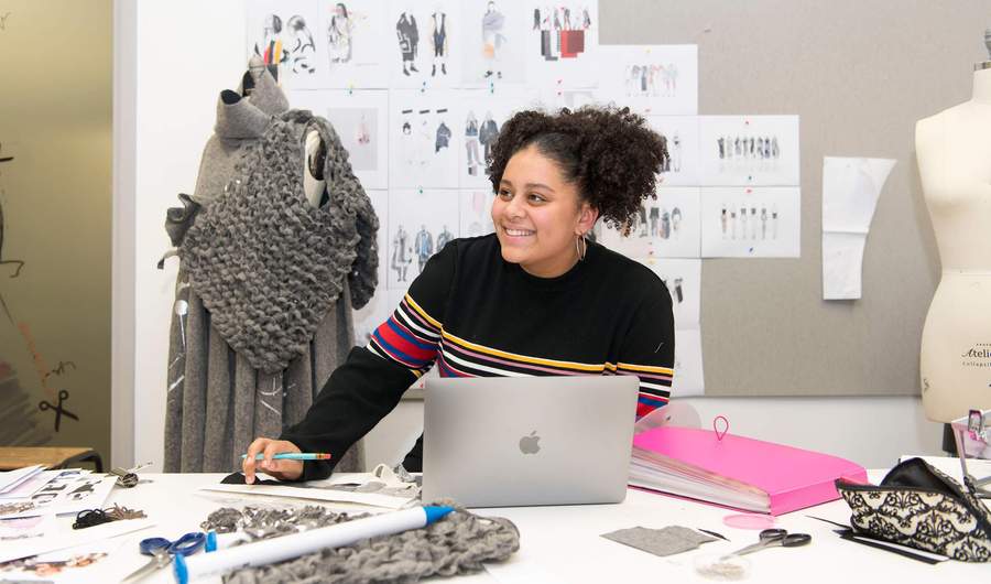 A young woman sits at a desk with a laptop computer and selection of fabric swatches. Behind her is a mannequin and wall of clothing designs.