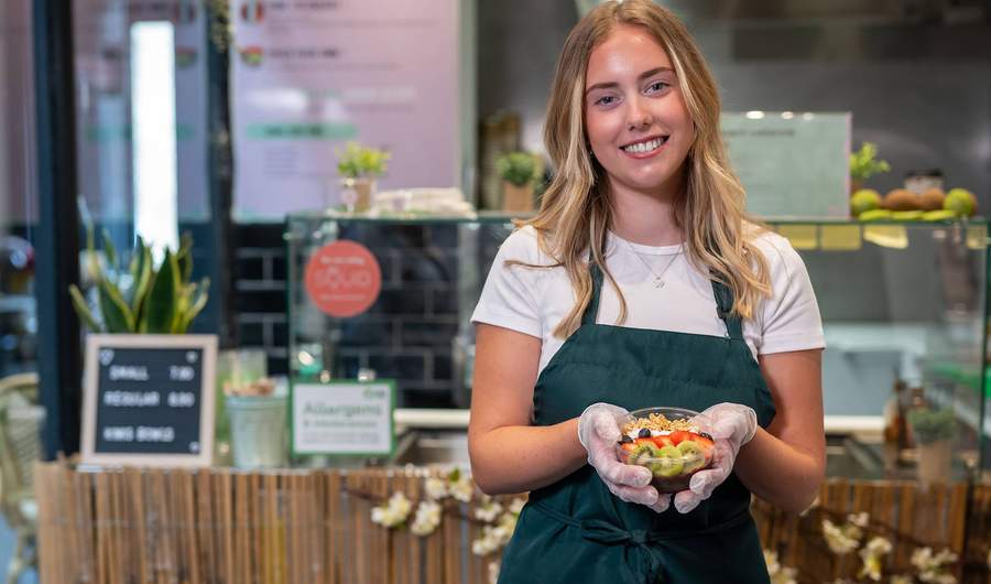 A young person wearing a green apron holds a fruit bowl while standing in front of a kitchen counter.