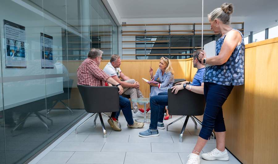 A group of people sit around a small table engaged in conversation. They are in a meeting space with a glass wall. A corridor is visible through the glass.