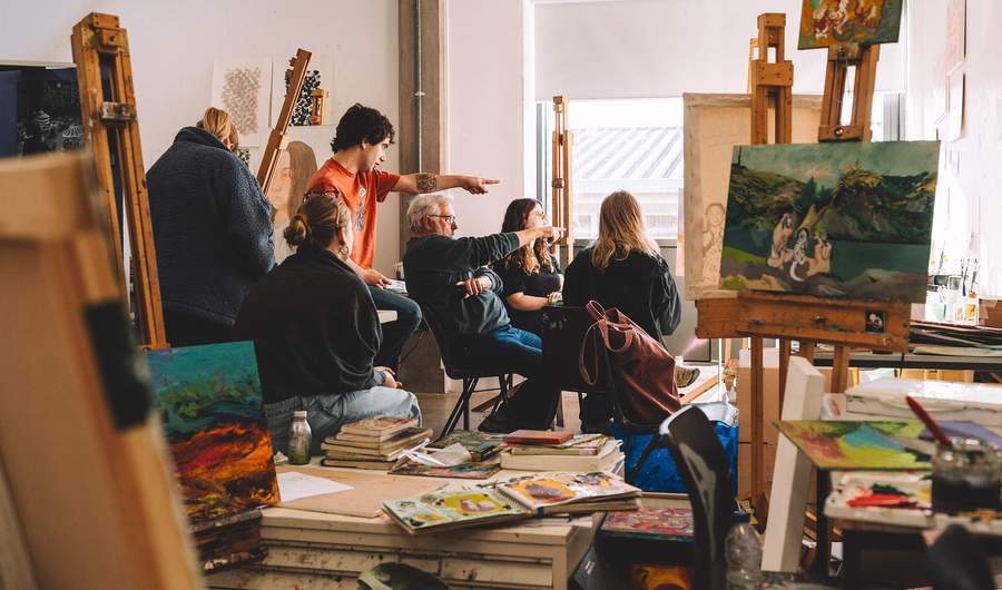A group of students study a canvas painting mounted on an easel. There are other canvasses on the tables around them, along with sketchbooks.