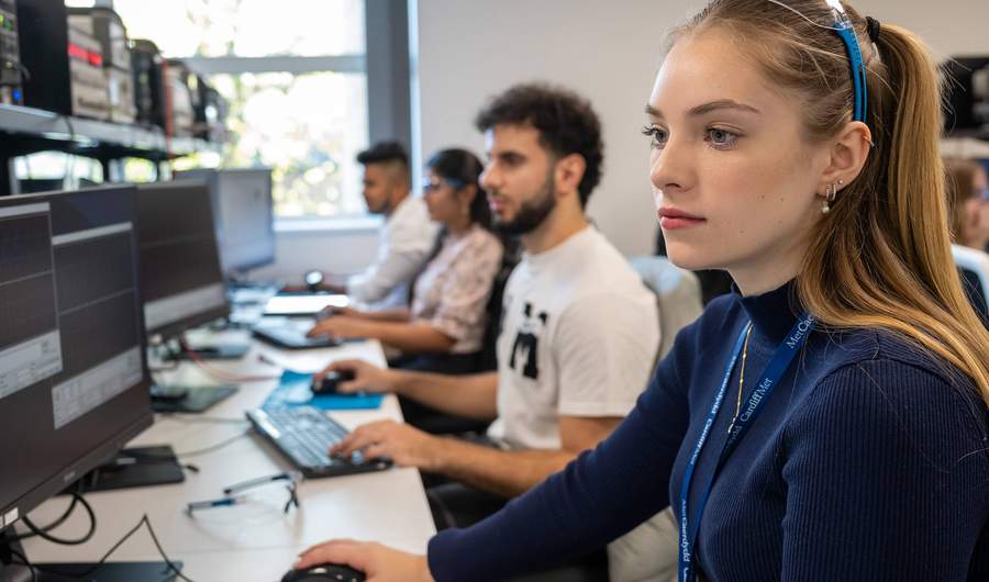 A group of people sit alongside each other at a long desk. Each of them is using a computer workstation.