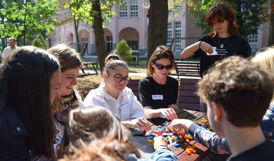 A group of people wearing name badges sit around a table outdoors. On the table in front of them are LEGO bricks.
