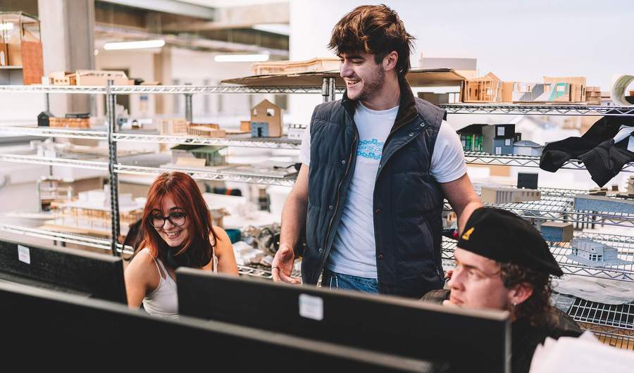 Students gathered around computer workstations in a bright studio space with architectural models displayed on shelves behind.