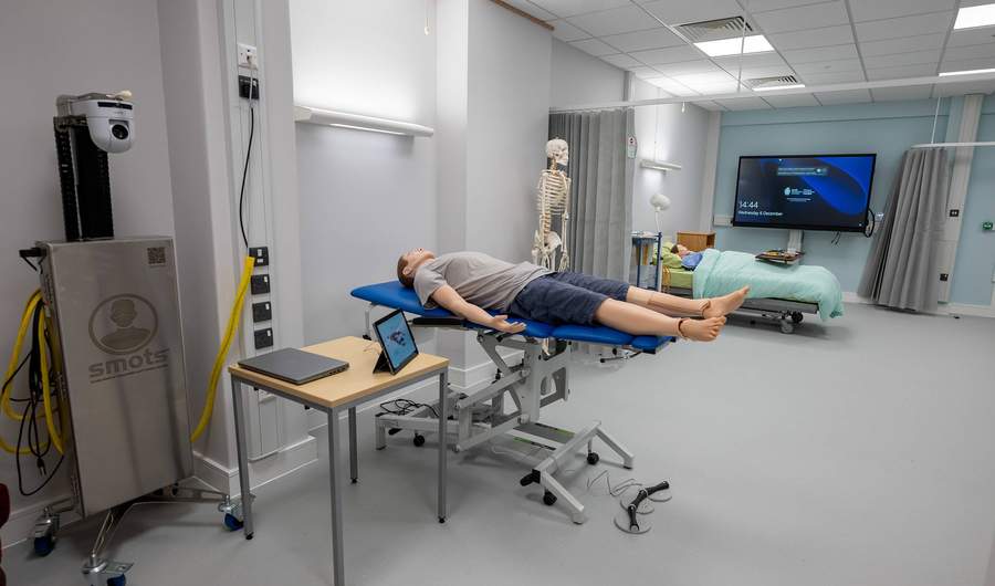 A manikin lies on a height-adjustable trolley in a simulated clinical environment.