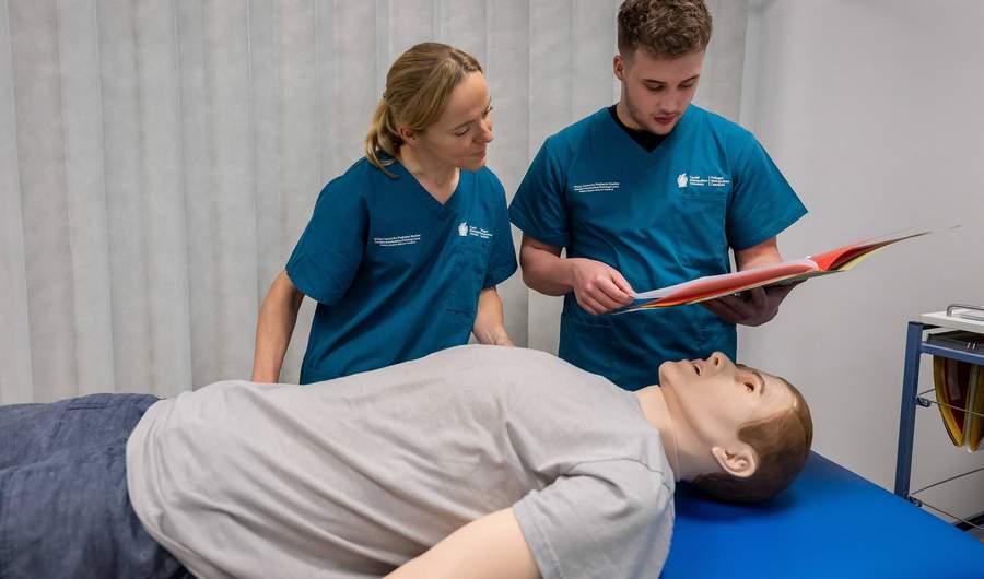 A manikin lies on a trolley in a simulated clinical environment, while a student consults patient records.