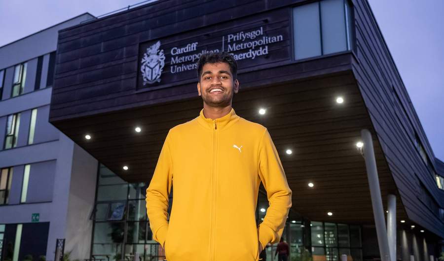 A young man in a yellow sweatshirt stands in front of a large modern building.