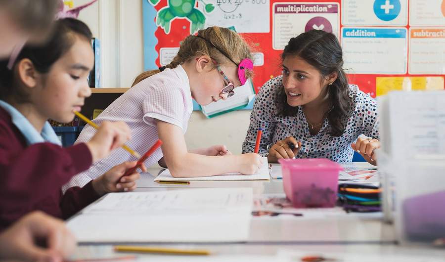 A school teacher sits at a table next to two young children. The children are writing in notebooks.