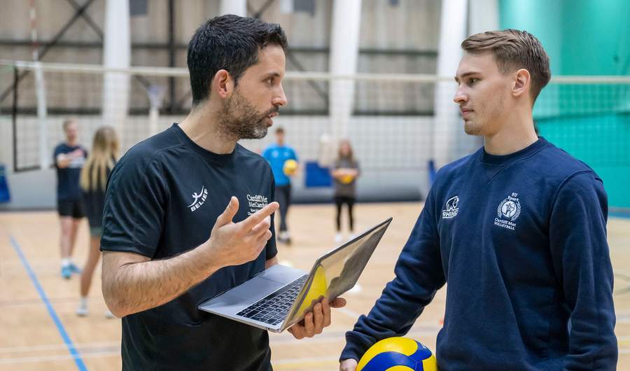 Two people converse in a gym. One holds a laptop computer, while the other holds a volleyball.