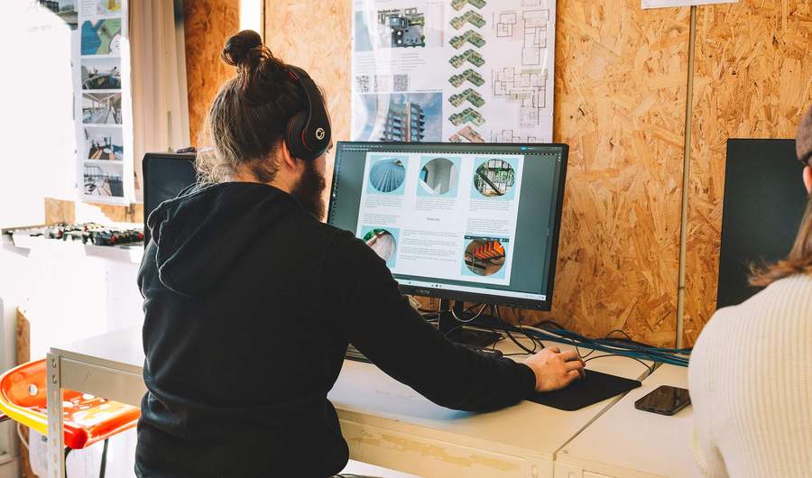 Student using a desktop computer at a workstation with architectural posters on the wall.