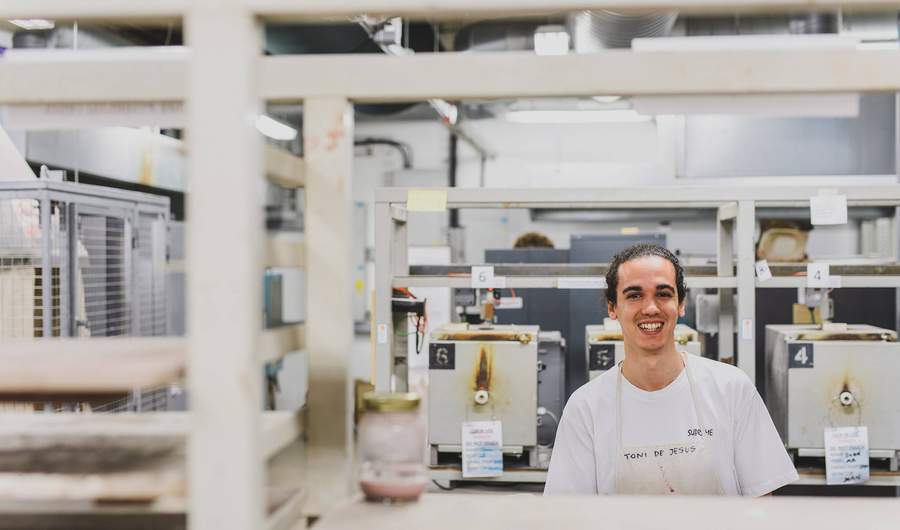 A young man stands in a ceramics workshop. Behind him is a row of small kilns.