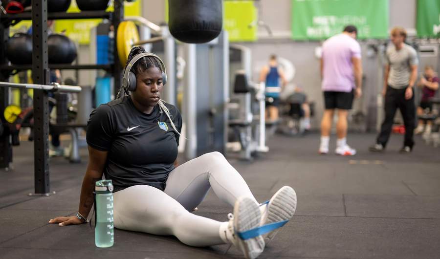 A woman sitting on the gym floor, engaged in a fitness activity, with weights and mats visible around her.