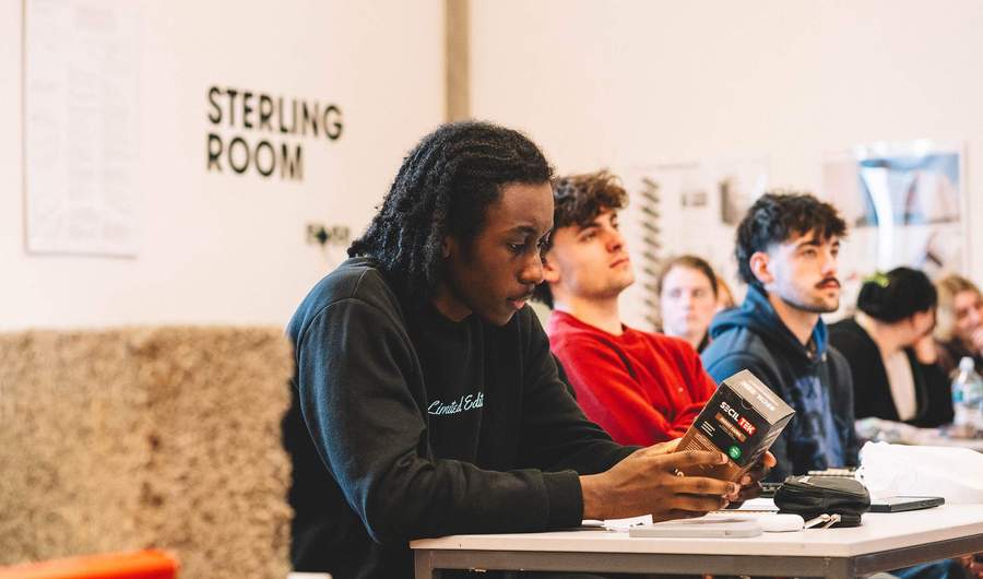 Student holding a boxed product while seated at a classroom table with others listening nearby.