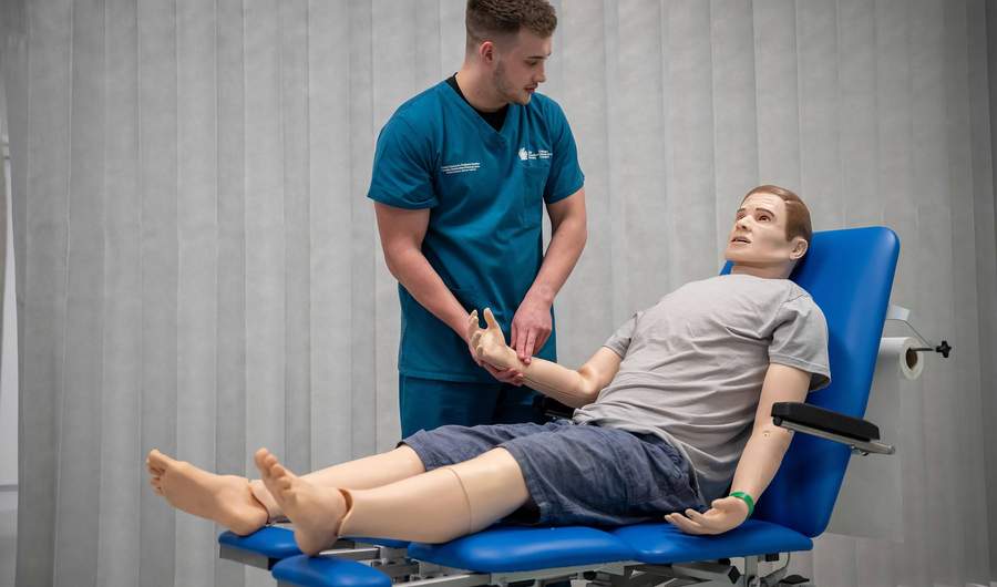 A student takes the pulse of a manikin lying on a height-adjustable trolley in a simulated clinical environment.