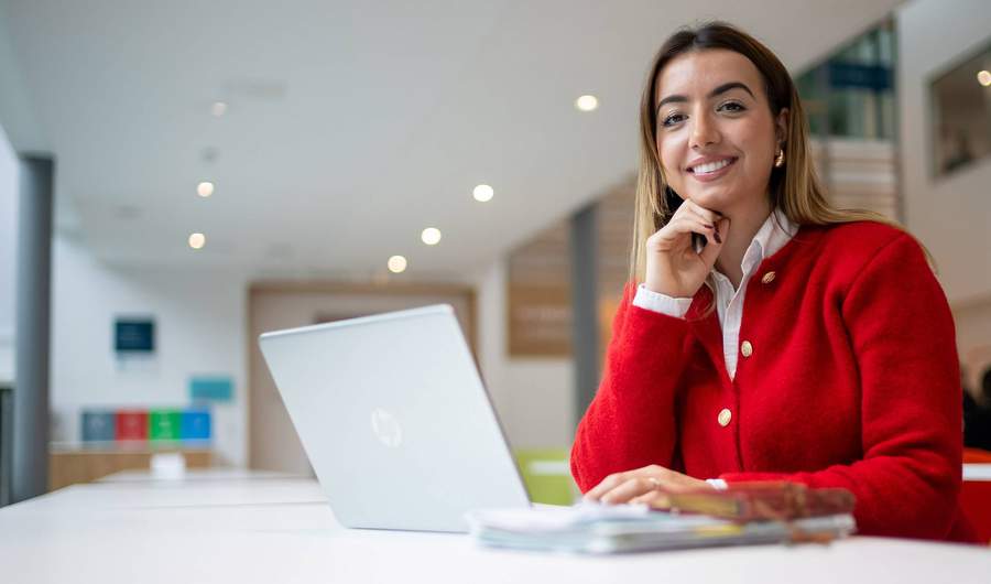 A person is smiling while sitting at a table with a laptop in front of them. They are in a bright, modern setting.