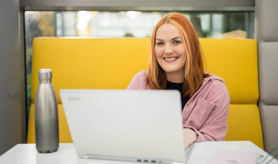 A young woman is smiling while sitting at a table with a laptop in front of them. They are in a bright, modern setting, with a yellow cushioned seating area behind her.