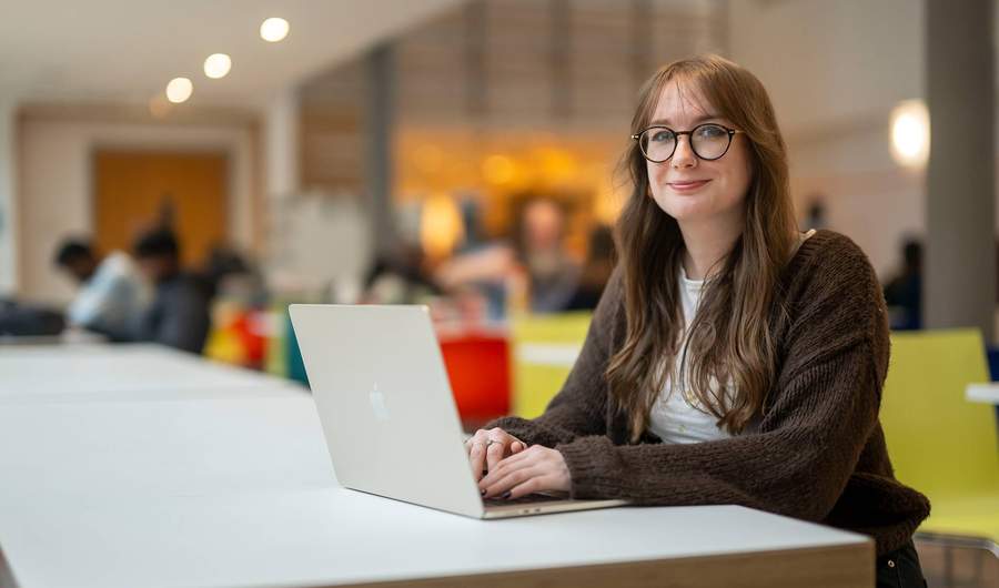 A person is smiling while sitting at a table with a laptop in front of them. They are in a bright, modern setting.