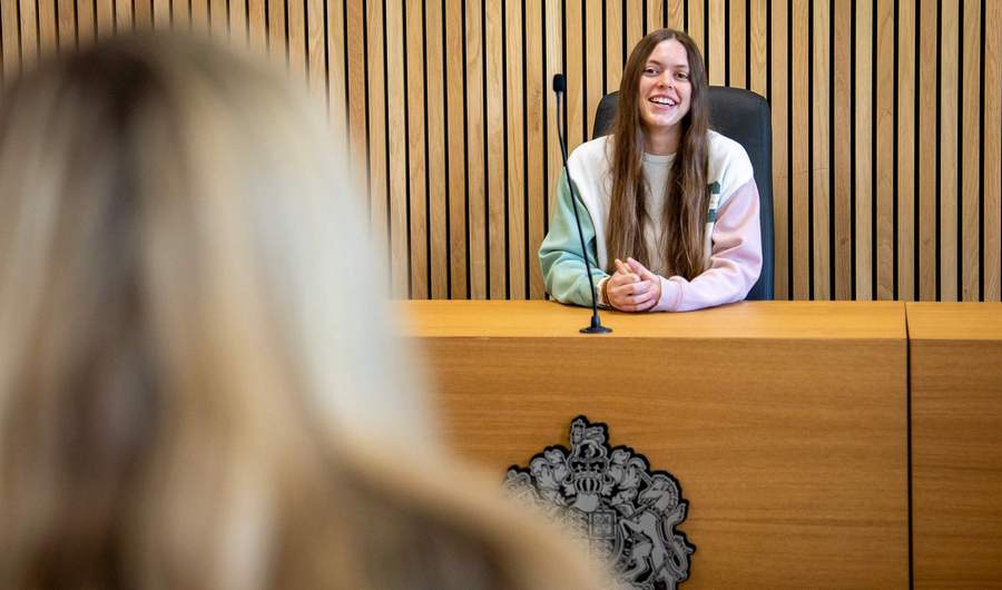 A person sits at a courtroom bench. On the front of the bench is a large crown seal.