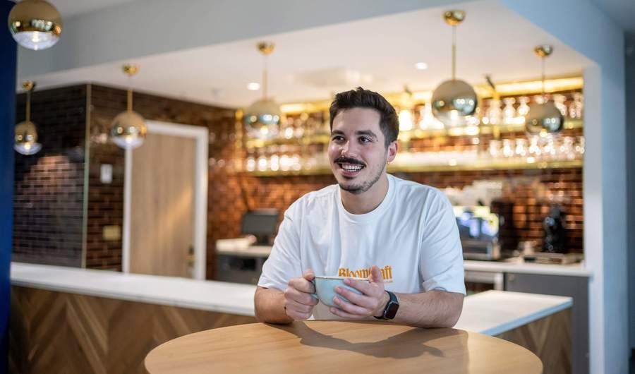 A young man sits at a table holding a coffee cup. Behind him is a bar area with racks of glasses.