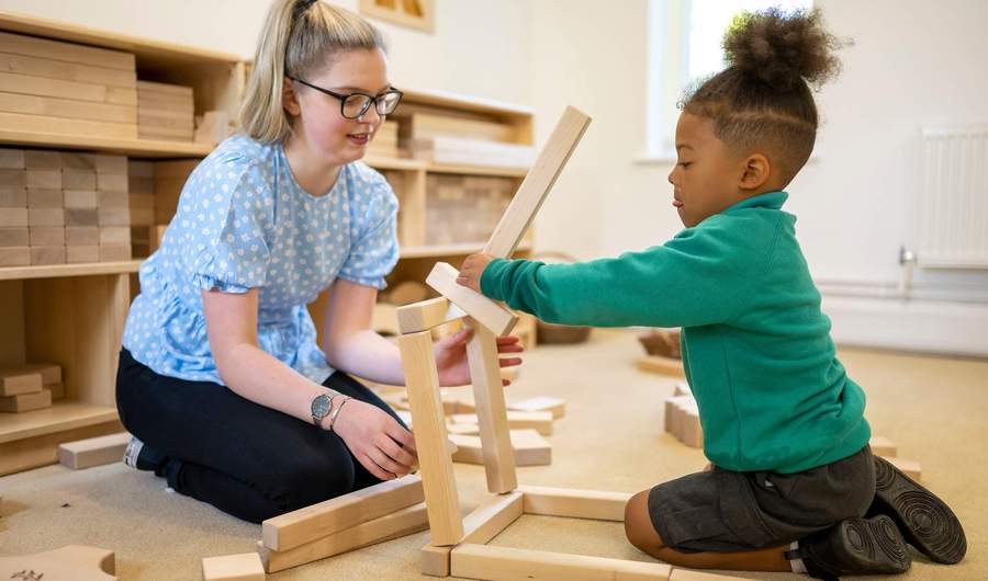 A child plays with wooden blocks as a teacher looks on.