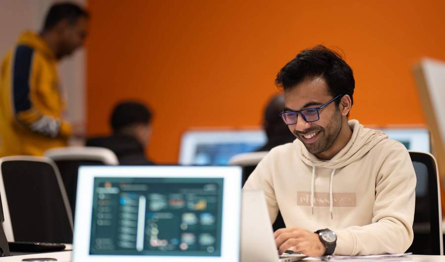 A student wearing a white hoodie and glasses is focused on his laptop while working at a desk.