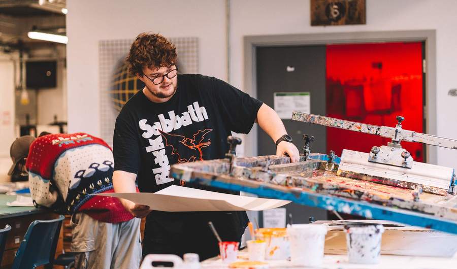 A student lifts a sheet of paper from underneath a printing press.