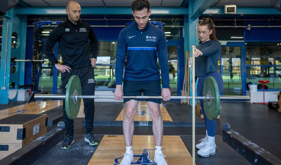 A person lifts a weight in a gym while two other people watch.