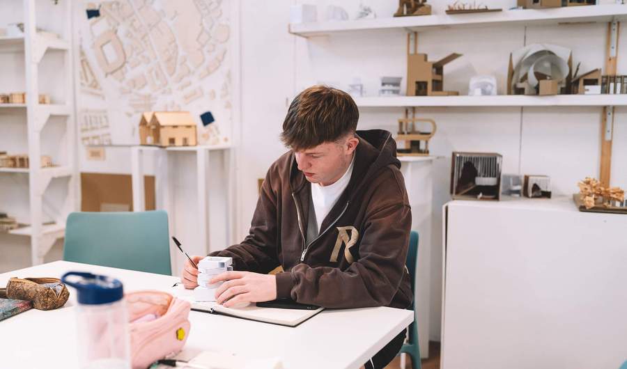 Student sitting at a desk writing in a notebook with architectural models displayed on shelves behind.