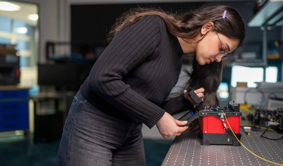 A student pressing the touchscreen of a small electronic device. In the background are other devices and computer monitors.
