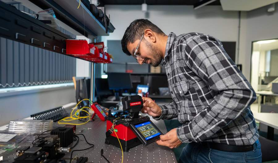 A student pressing the touchscreen of a small electronic device. In the background are other devices and computer monitors.