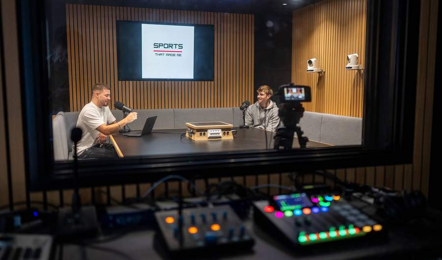 Two people sit at a table in a sound booth talking to one another. In front of them are microphones and other audio equipment.