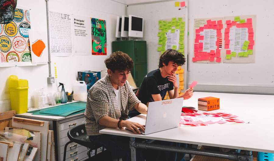 Two students sit at a table. One uses a laptop while the other reads a Post It note. There are Post It notes on the table and on the walls.
