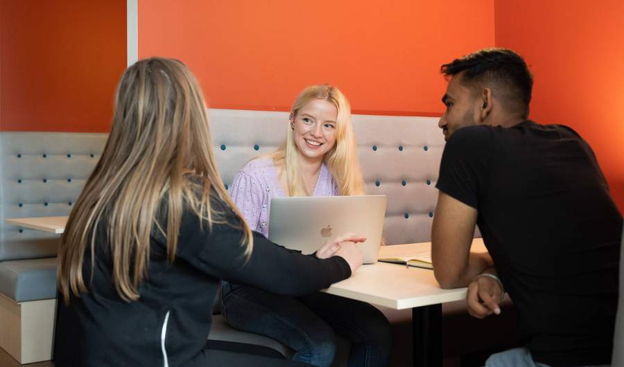 Three students sit together on bench seating at a table. The wall behind them is a vibrant orange red.
