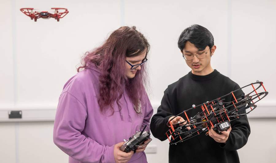 A person holding a drone quadcopter points to one section of it. A second person holding a remote control looks on. In the background a smaller quadcopter hovers in the air.