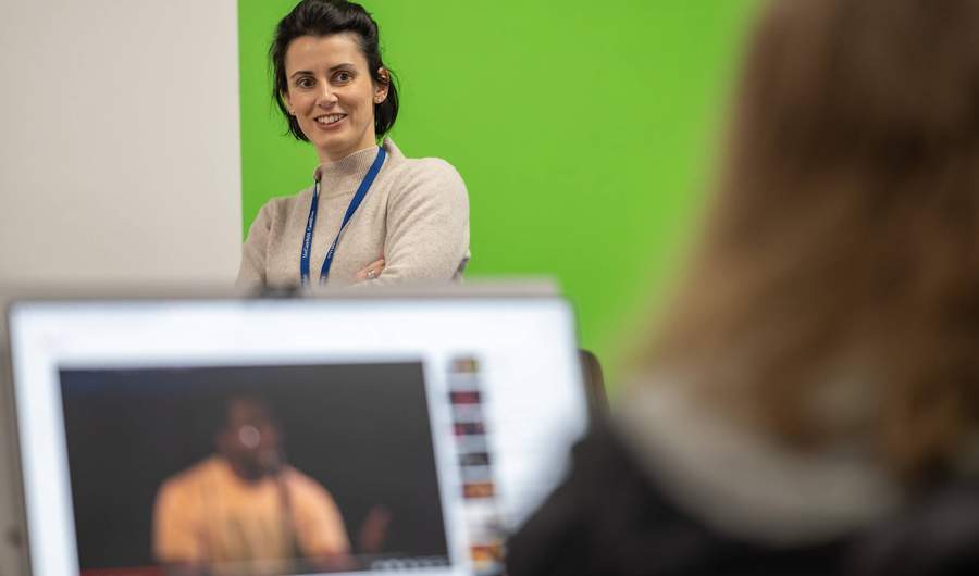 A person stands with their arms folded. Facing them is another person with an open laptop.