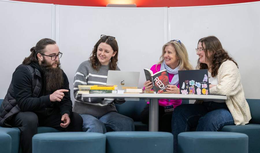 A group of people sit around a circular table. On the table are several books.