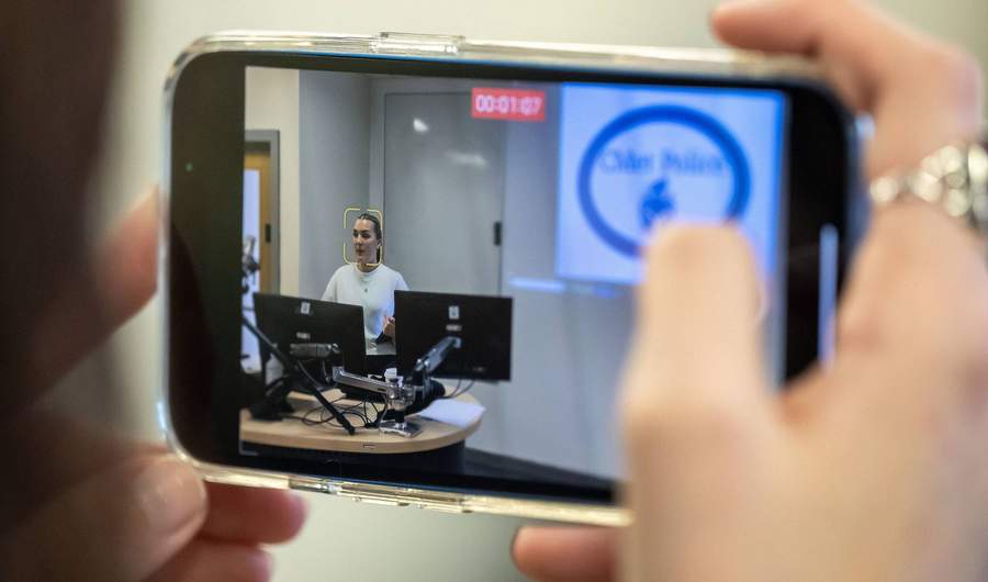 A close-up of a smartphone screen recording a video. On the screen is a person sat at a desk in front of two computer monitors.