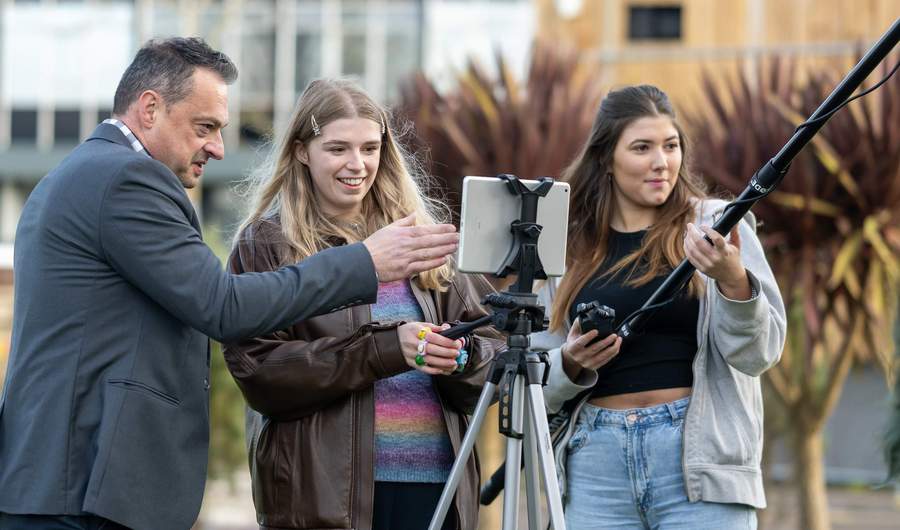 A group of people film outside. One person is operating a camera, while another holds a microphone boom.