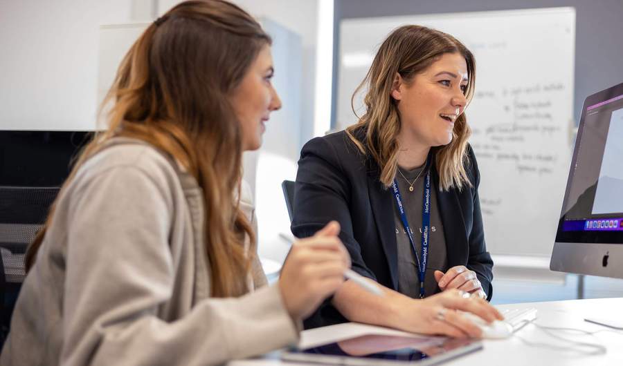 Two people sat at a desk in front of a Mac computer.