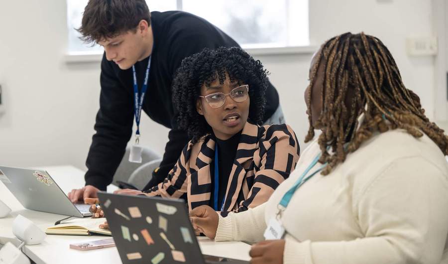 Three people working alongside each other at a desk. In front of them are two laptop computers.