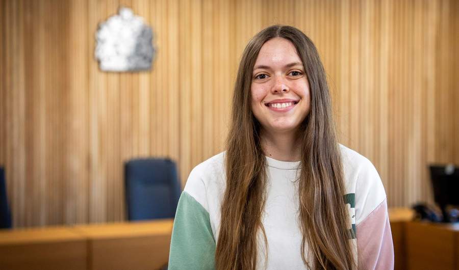 A person stands in a courtroom. In the background is a bench with a crown seal above it.