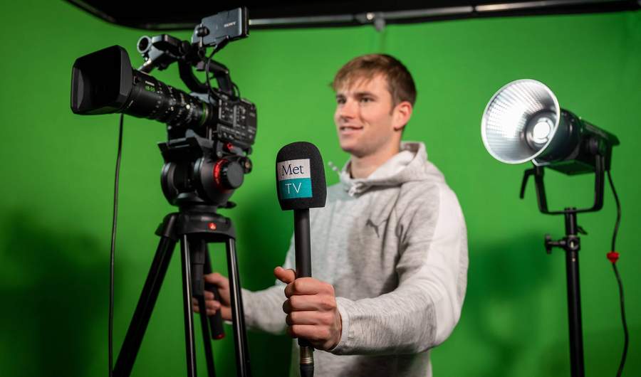 A person stands behind a television camera. Behind them is a green backdrop.