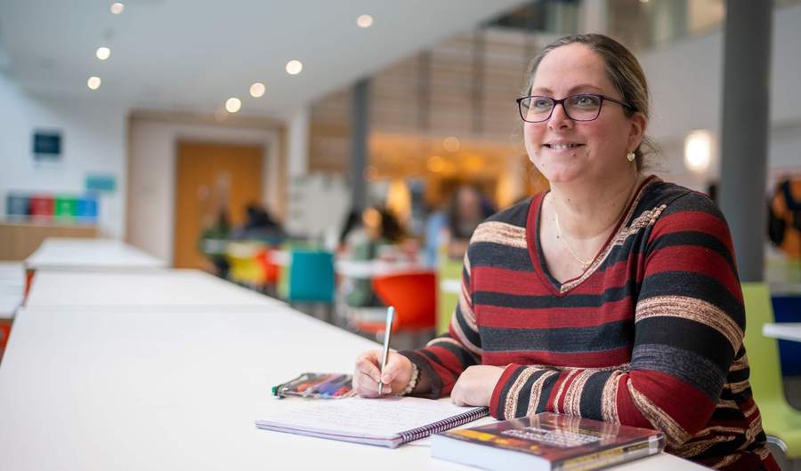 A person sits a long table writing in a notebook. In the background are other people sat at smaller tables.